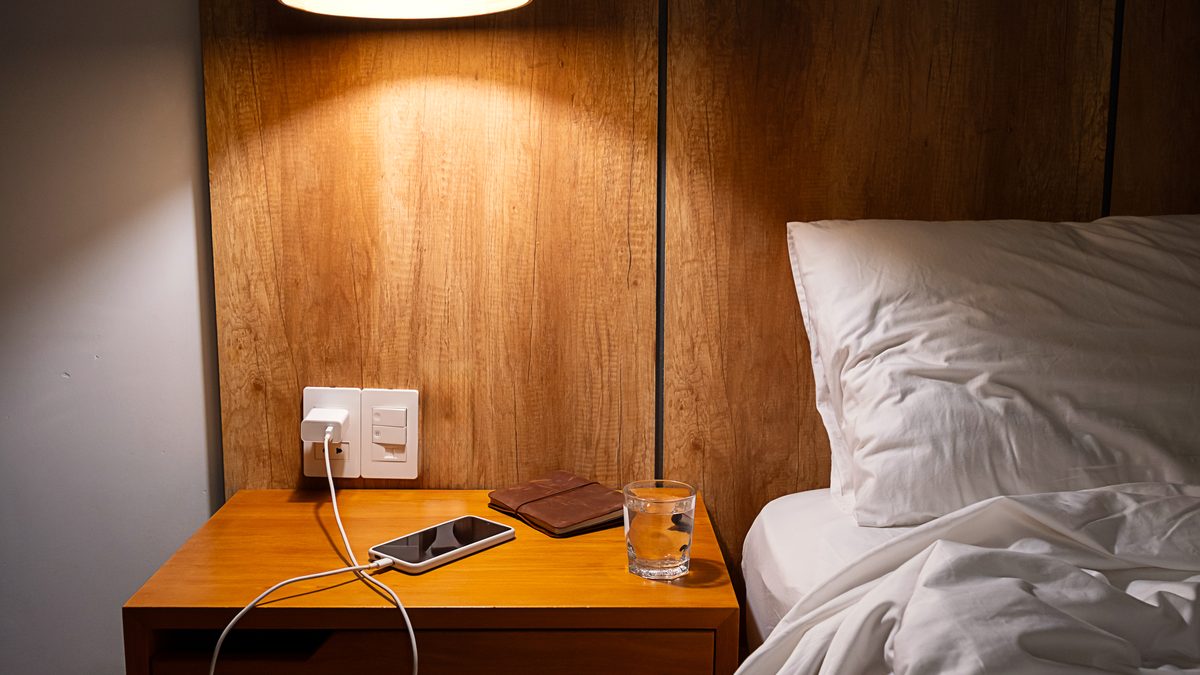 Ilustrační stock: telefon nabíjí na nočním stolku vedle lampy, postel v pozadí, civilně, bez loga